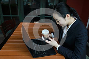 Young woman sitting in coffee shop at wooden table, drinking coffee and using smartphone.On table is laptop