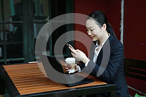 Young woman sitting in coffee shop at wooden table, drinking coffee and using smartphone.On table is laptop