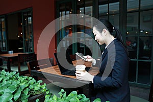 Young woman sitting in coffee shop at wooden table, drinking coffee and using smartphone. On table is laptop