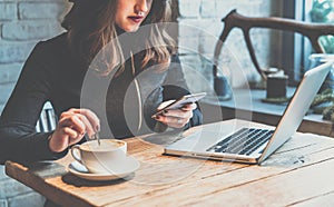 Young woman sitting in coffee shop at wooden table, drinking coffee and using smartphone.On table is laptop.