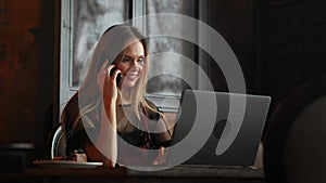 Young woman sitting in coffee shop at wooden table, drinking coffee and using smartphone.On table is laptop. Girl