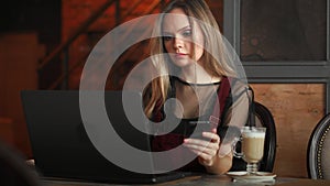 Young woman sitting in coffee shop at wooden table, drinking coffee and using smartphone.On table is laptop. Girl