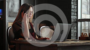 Young woman sitting in coffee shop at wooden table, drinking coffee and using smartphone.On table is laptop. Girl