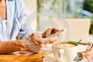 Young woman sitting in coffee shop at wooden table, drinking coffee and using smartphone.On table is laptop. Girl