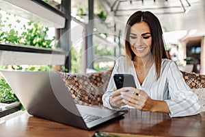 Young woman sitting in coffee shop at wooden table, drinking coffee and using smartphone on table is laptop. Girl browsing