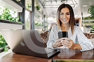 Young woman sitting in coffee shop at wooden table, drinking coffee and using smartphone on table is laptop. Girl browsing