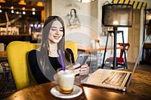 Young woman sitting in coffee shop at wooden table, drinking coffee and using smartphone. On table is laptop. Girl browsing intern