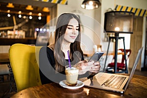 Young woman sitting in coffee shop at wooden table, drinking coffee and using smartphone. On table is laptop. Girl browsing intern