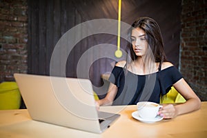 Young woman sitting in coffee shop at wooden table, drinking coffee and using smartphone. On table is laptop. Girl browsing intern