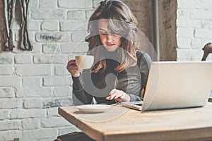 Young woman sitting in coffee shop at wooden table, drinking coffee and using smartphone.On table is laptop. Girl browsing interne