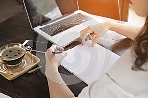 Young woman sitting in coffee shop at wooden table, drinking coffee and using smartphone.On table is laptop for internet shopping