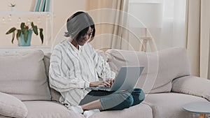 Young woman sitting on comfortable sofa in living room typing on laptop female freelancer working remote at home pretty