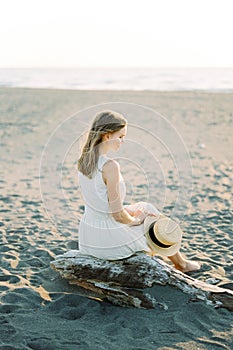 Young woman sitting on a driftwood on the beach with a straw hat in her hands