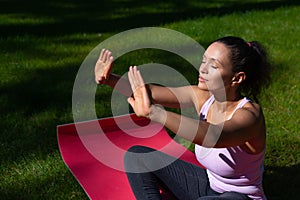 Young woman sitting on a fitness mat on a green grass in the park enjoying sunny day while performing yoga