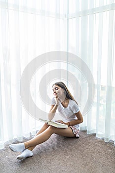 Young woman sitting on a floor near a window with a book. Young girl reading at home