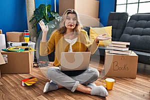 Young woman sitting on the floor at new home using laptop clueless and confused expression with arms and hands raised