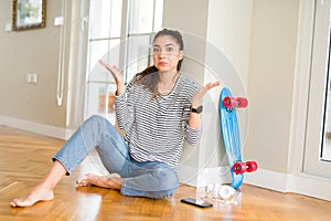 Young woman sitting on the floor using skateboard and headphones clueless and confused expression with arms and hands raised