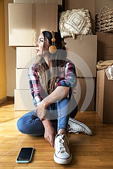 Young woman sitting in front of pile of cardboard boxes wearing headphones. Pensive female looking out the window