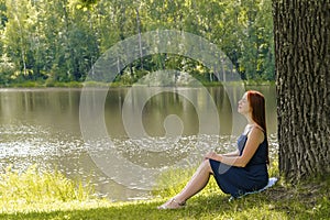 Young woman sitting on grass under high big oak,trunk tree near lake,river,meditating,relaxing in National Park.Teen girl,