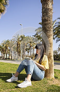 Young woman sitting on grass and writing in a notebook outside surrounding with palm trees.