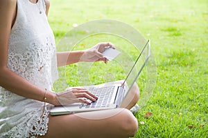 Young woman sitting on green grass in park. female using laptop, holding credit card typing on notebook. online shopping concept.