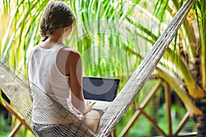 Young woman sitting in a hammock with laptop in a tropical resort. back view.