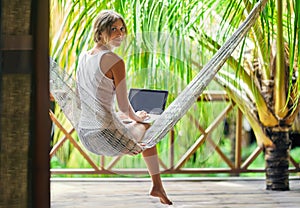 Young woman sitting in a hammock with laptop in a tropical resort. back view.