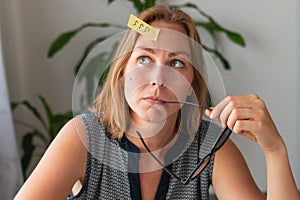 A young woman sitting at her Desk, holding glasses near mouth and thinking about something. On the forehead sticker with question