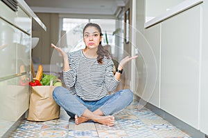 Young woman sitting on the kitchen floor with a paper bag full of fresh groceries clueless and confused expression with arms and