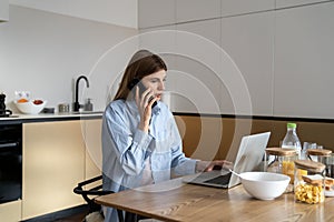 Young woman sitting at kitchen table using laptop computer and talking by phone with client