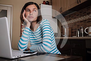 Young woman sitting in kitchen and working on laptop in morning. Female using laptop dining table