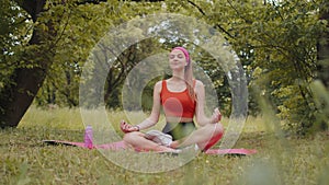 Young woman sitting on mat in lotus position, relaxing, practicing yoga meditation in city park