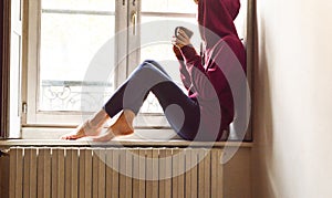 Young woman sitting near the window looking outside drinking coffee in a nostalgic mood