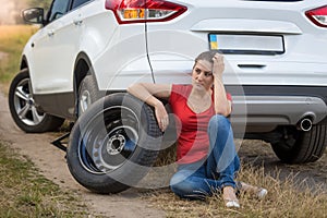 Young woman sitting next to car with flat tire in field and waiting for help