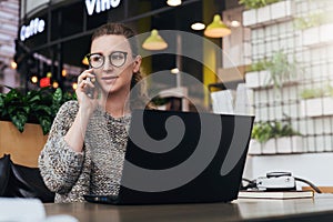 Young woman sitting in office, cafe at table in front of laptop and talking on mobile phone. Freelancer works in a cafe.