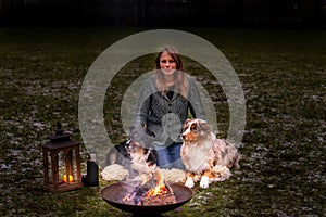 Young woman is sitting outside in the woods with her two Australian Shepherd dogs. Snow on the grass, at night by the campfire.