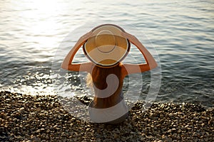 Young woman is sitting on the pebble shore with beach dress on holding a straw hat on her head. Back view portrait of