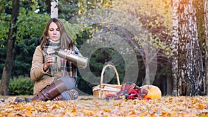 Young woman sitting on picnic pouring hot tea from a thermos in autumn park. Girl sits the rug near the pumpkin