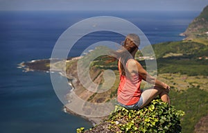 Young woman sitting on a precipice, view from behind. Incredible view of small village on ocean coast