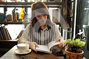 Young woman sitting in restaurant and reading book with cup of c