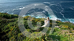 Young Woman Sitting On The Rock In The Hills With Wildflowers And Admiring The Blue Ocean