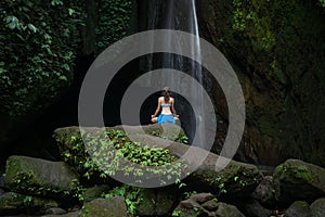 Young woman sitting on the rock, practicing yoga near waterfall. Hands in gyan mudra. Leke Leke waterfall, Bali. View from back