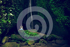 Young woman sitting on the rock, practicing yoga near waterfall. Hands in gyan mudra. Leke Leke waterfall, Bali. View from back