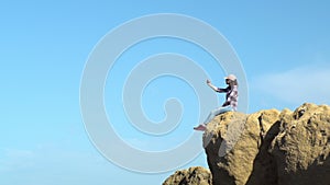A young woman is sitting on a rock with a telephone. Makes a selfie on a rock. A woman in a cap dangled her legs over a