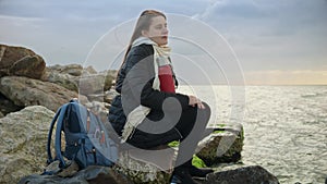 Young woman sitting on a rocky coastline, holding a thermos of hot tea as she gazes out at the stormy ocean.