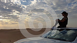Young woman sitting on the roof of a car and looking on the epic sunset, against the background of the desert.