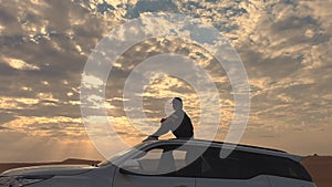 Young woman sitting on the roof of a car and looking on the epic sunset, against the background of the desert.