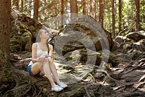 Young woman sitting on the roots of trees in wild forest. Fairy-tale forest and modern snow white