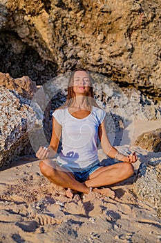 Young woman sitting on the sand near the rock. Hands in gyan mudra. Lotus pose. Yoga and meditation concept. Caucasian woman