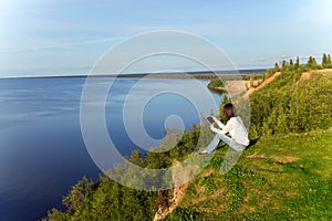 Young woman sitting on the shore of the lake with tablet compute